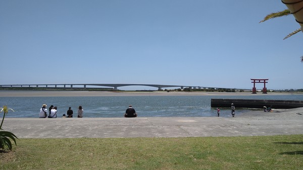 Den röda toriin står på ett rev innanför sundet mellan Hamanako och havet. I bakgrunden Hamanako-bron som går över sundet. Sanden är grå och vattnet strömt. I förgrunden en plattform av betong med samma färg som sanden.