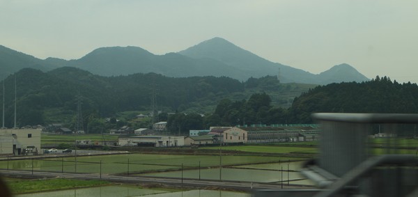 Den japanska landsbygden sedd från ett snabbtåg är lite enformig, men formen är roligare än städernas. Skogar identiska med kullar och berg tornar över små och stora samhällen med oändligt varierade småhus, småskaliga jordbruk och enstaka industrier som denna. Fabriken heter ＪＡさが みどり地区嬉野大型製茶工場. Foto taget från Kamome-tåg på Kyūshū.
